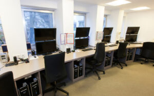Schaltzentrale, Einzelplätze an einem großen Schreibtisch mit zwei Monitoren, Stühle und Rechner